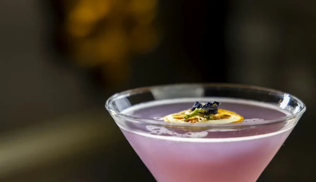 Frothy lavender cocktail in martini glass with dried orange and flowers.