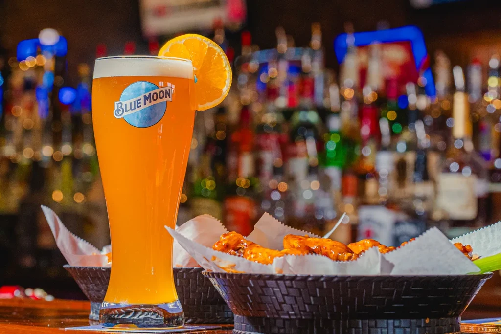 Blue Moon beer with orange slice and sauced chicken wings at a bar.