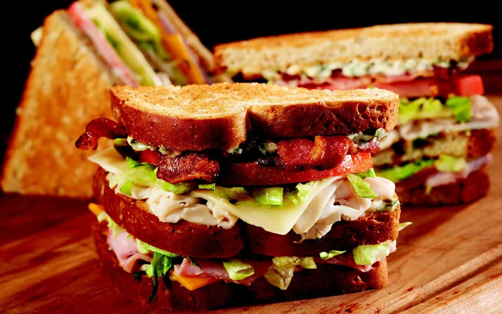Three large, layered sandwiches with meat, cheese, vegetables on wooden board.