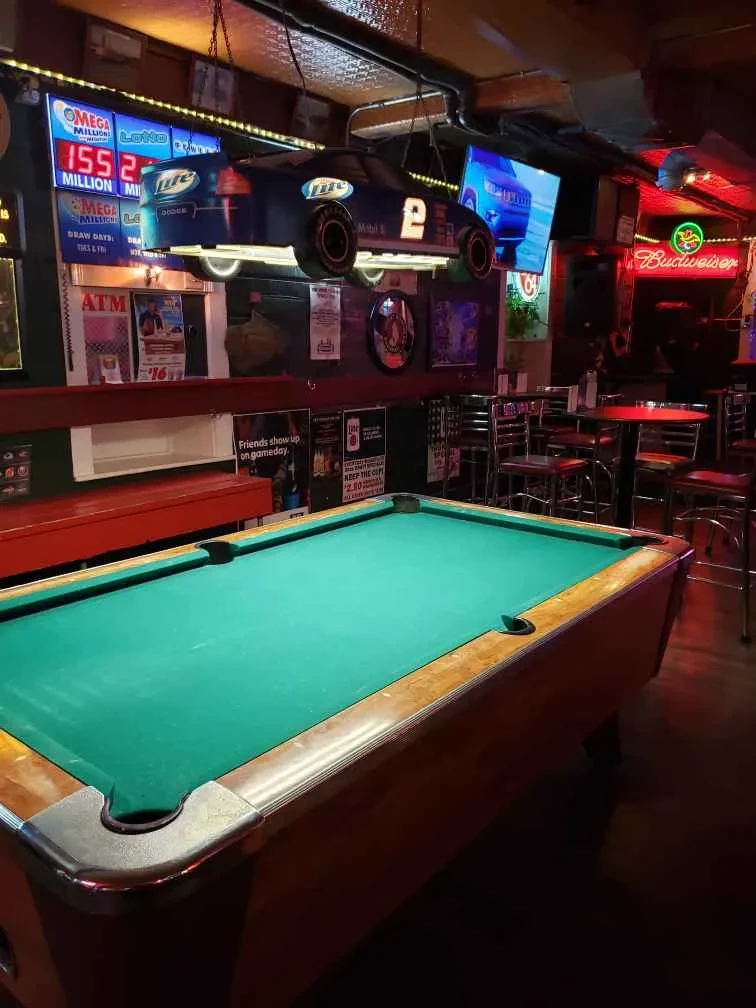 Dim bar interior, green pool table, neon signs, suspended race car.