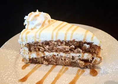 Slice of multi-layered carrot cake with cream cheese frosting, caramel, whipped cream.