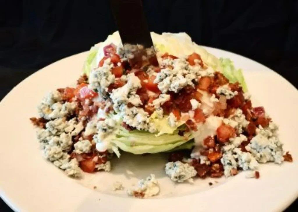 Iceberg wedge salad with blue cheese, bacon, tomatoes, dressing, and knife.