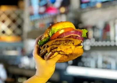 Hand holding a double-patty hamburger with cheese, lettuce, tomato, and onion.