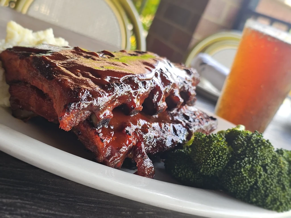 Plate of BBQ ribs, mashed potatoes, broccoli, and amber drink, outdoors.