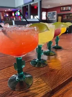 Five vibrant margaritas in green cactus glasses on a wooden bar.