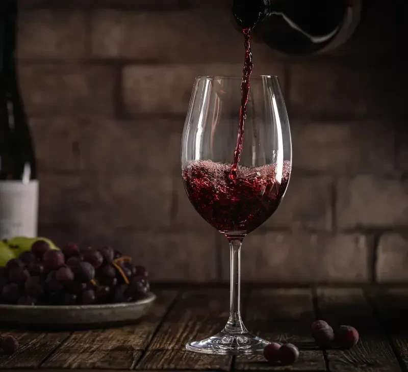 Red wine pours into a glass on a rustic table with grapes.