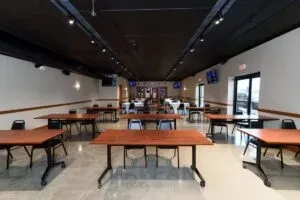 Empty event hall with wooden tables, black ceiling, and flat-screen TVs.
