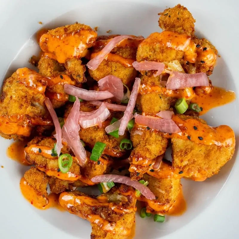 Fried cauliflower with orange-red sauce, sesame seeds, pickled onion, and green onions.