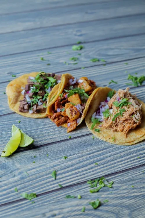 Three tacos with meat, potato, lime, cilantro on a wooden surface.