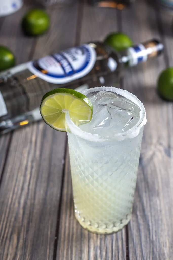 Salt-rimmed margarita with lime and tequila bottle on wooden table.