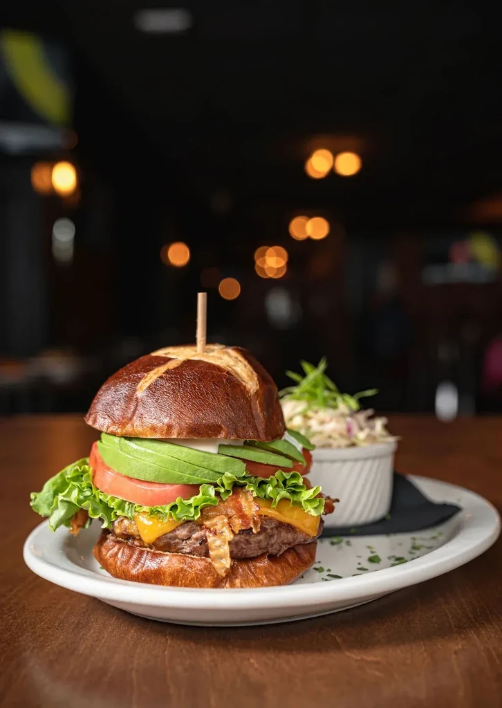 Pretzel bun burger with bacon, avocado, and coleslaw on plate.