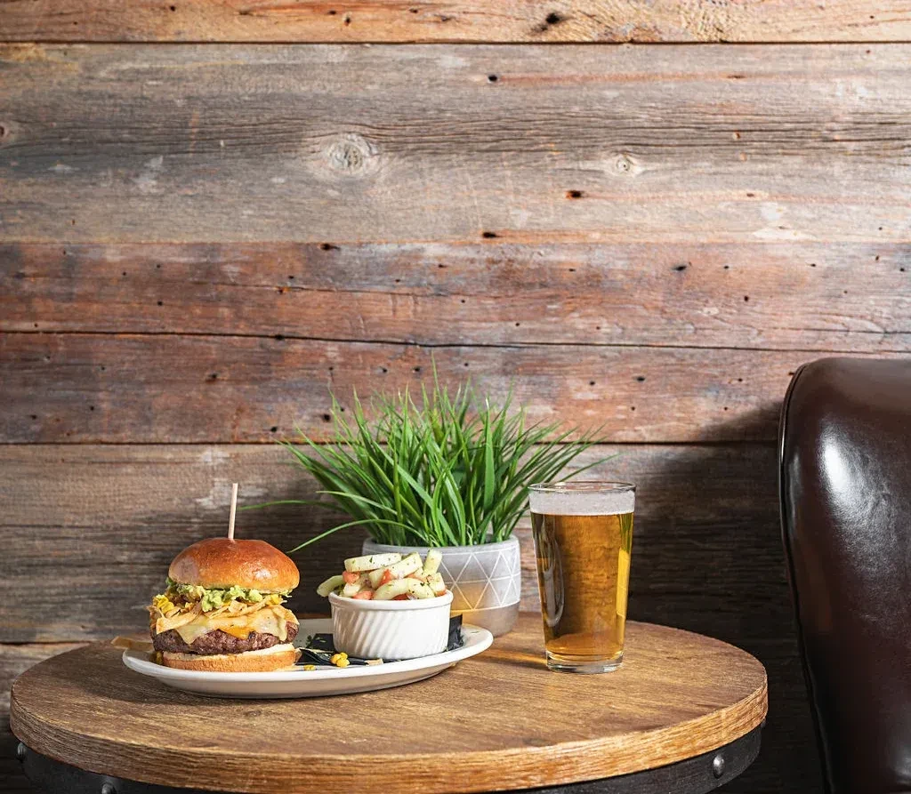 Gourmet burger, side dish, and pint of amber beer on wooden table.
