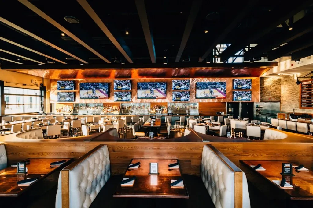 Modern sports bar interior: long bar, six TVs showing basketball, booths.