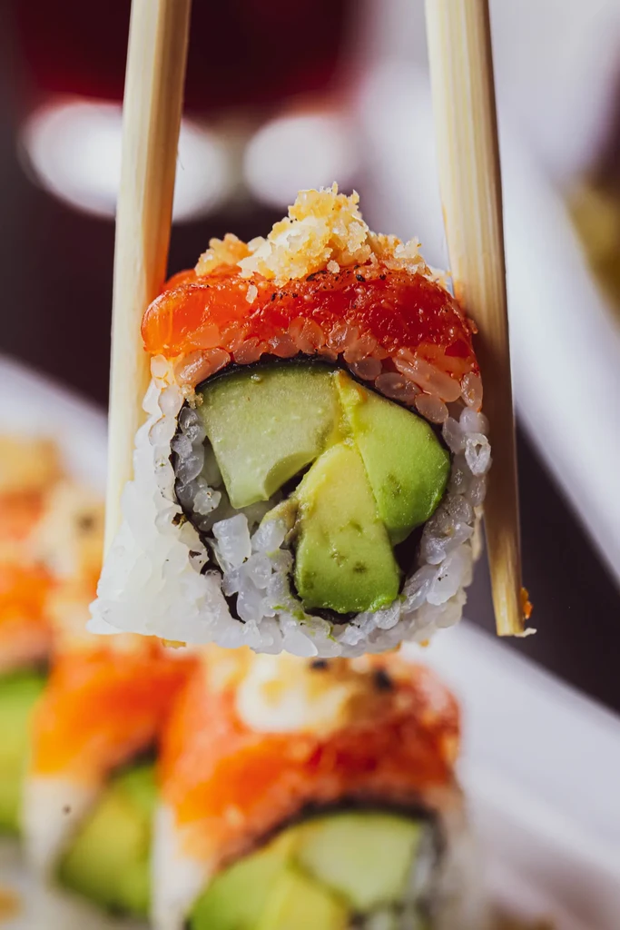 Chopsticks holding a sushi roll with avocado, salmon, and crispy flakes.
