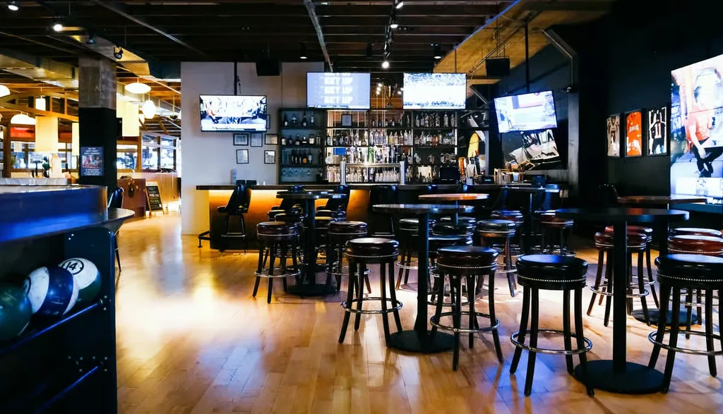 Modern bowling alley lounge featuring a stocked bar, TVs, and tables.