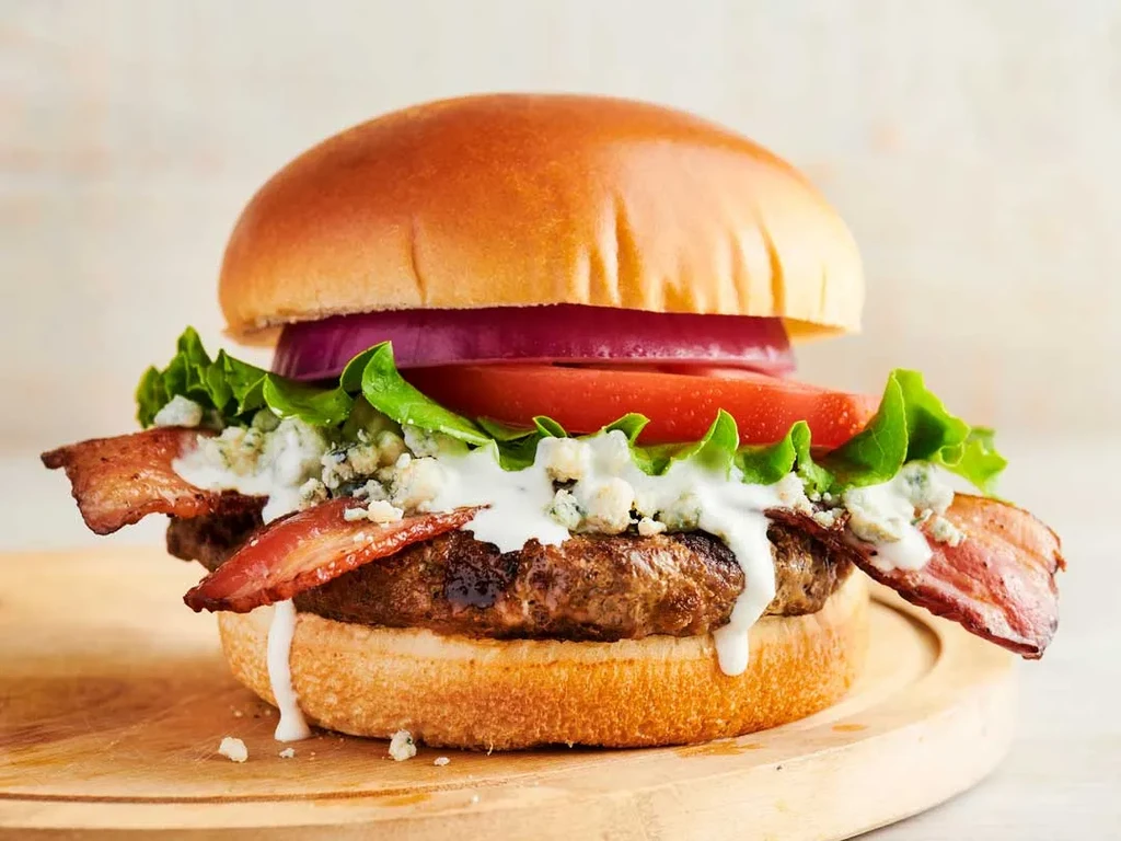 Gourmet burger with beef patty, bacon, blue cheese sauce, and vegetables.