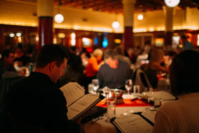 Two people read menus at a busy, warmly lit restaurant.