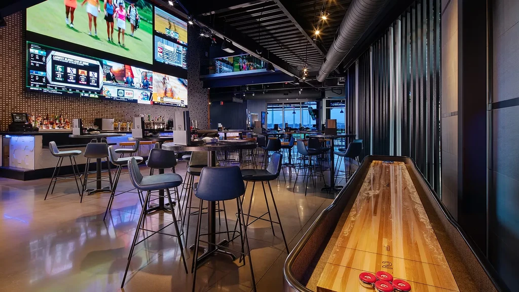 Modern sports venue with shuffleboard, blue-lit bar, and massive video wall.