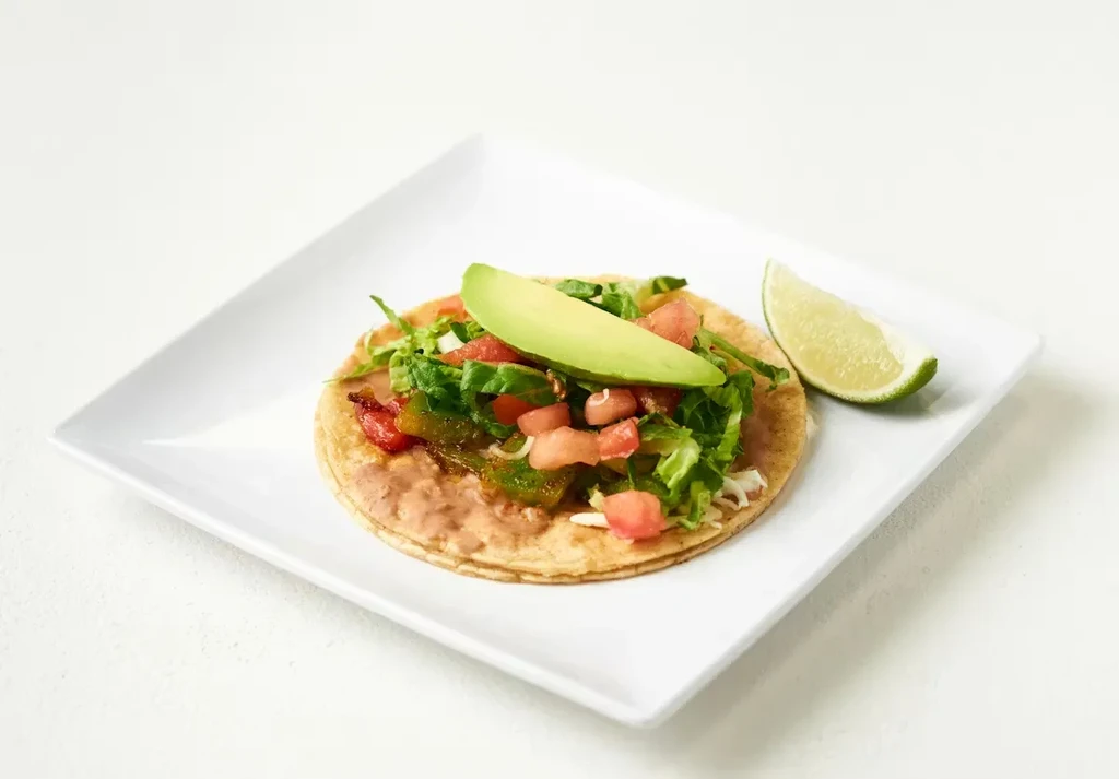 Tostada topped with beans, lettuce, tomatoes, avocado, and a lime wedge.