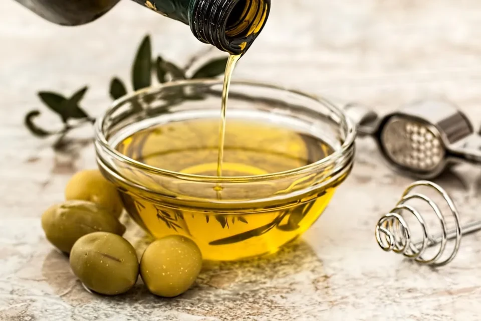 Olive oil pouring into a bowl with green olives and olive leaves.