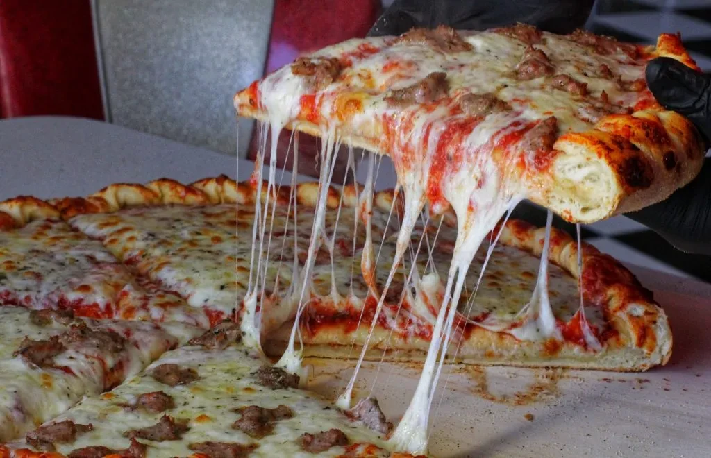 Gloved hand lifts sausage pizza, showing long strands of melted cheese.