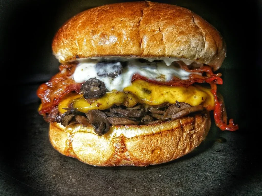 Close-up: Gourmet burger with melted cheese, mushrooms, bacon, creamy sauce.