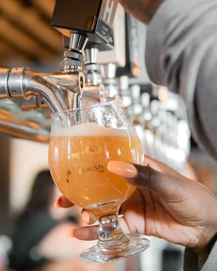 Hand holding a "PECKISH PIG" glass being filled with amber beer.