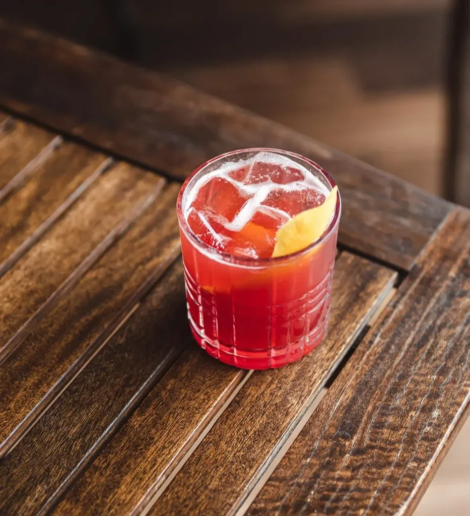 Red cocktail with ice and lemon wedge on a wooden table.