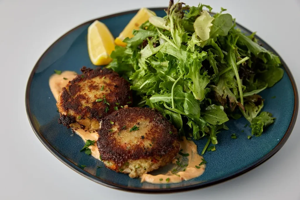 Two crab cakes, salad, and lemon wedges on a blue plate.