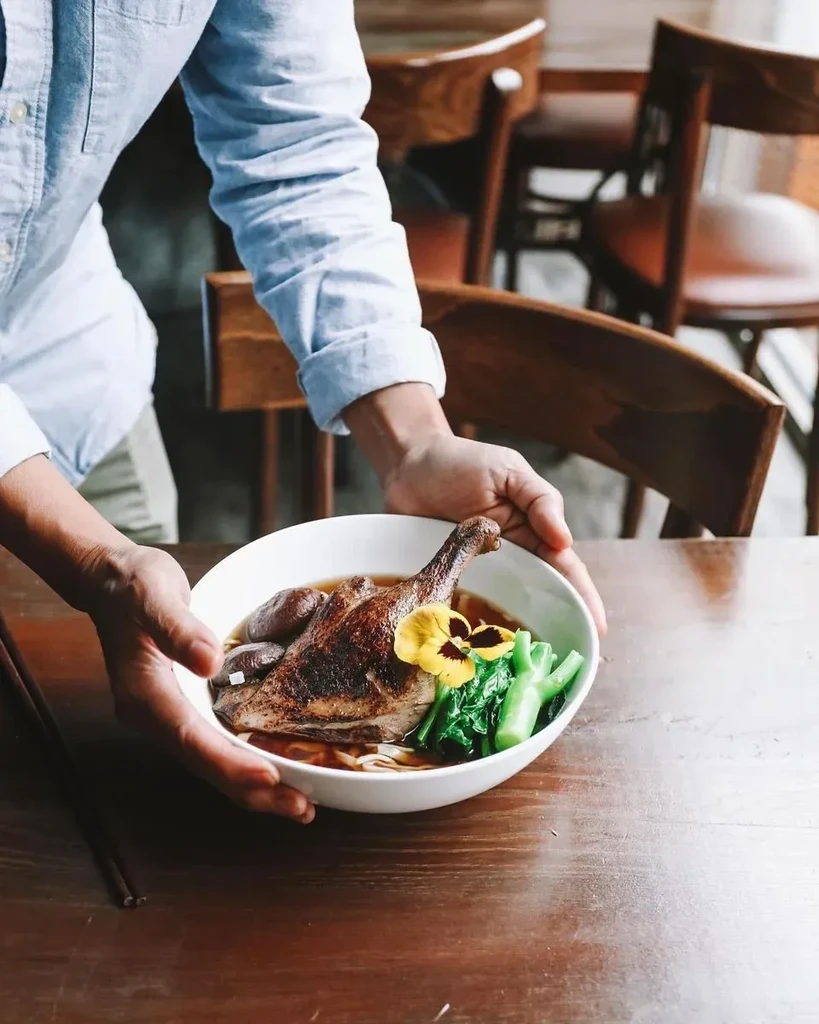 Hands holding a white bowl of duck noodle soup.
