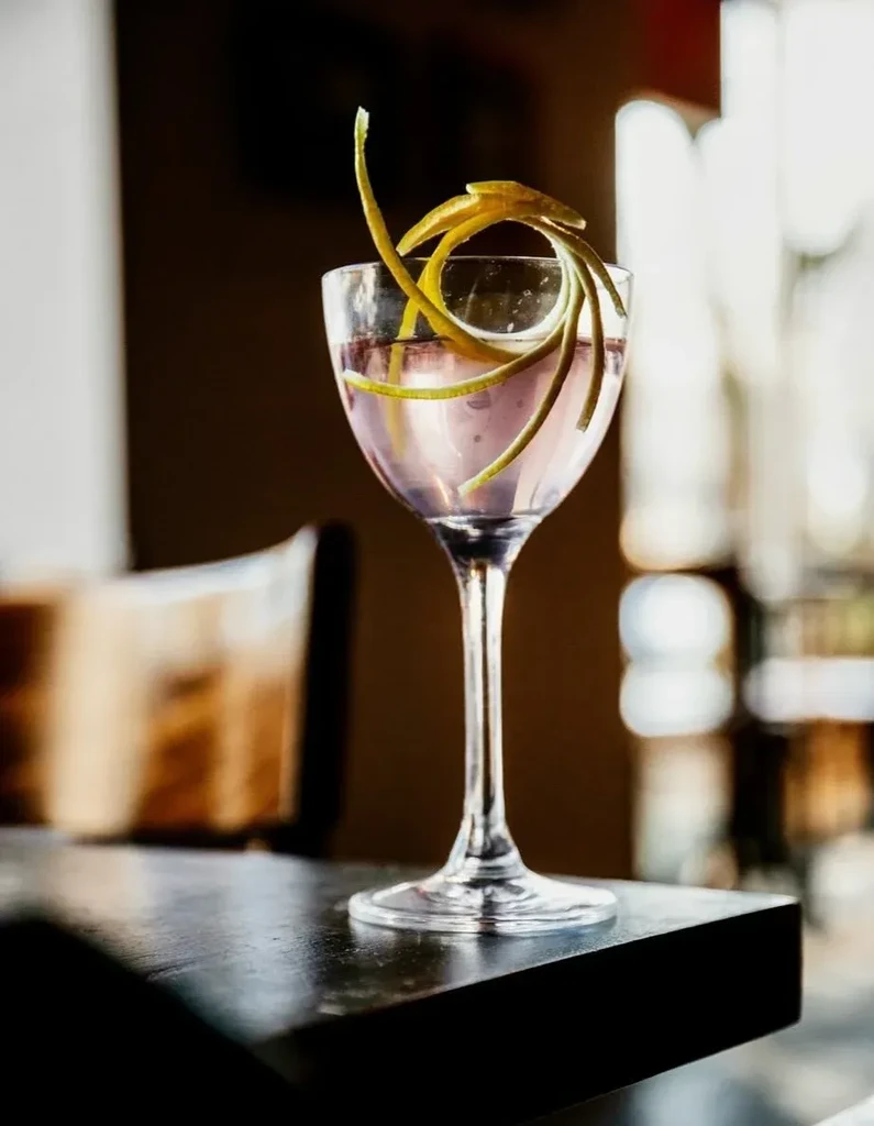 Cocktail in coupe glass, garnished with a citrus peel, on dark surface.