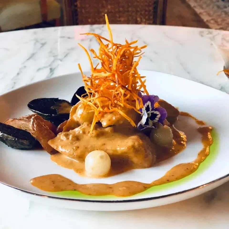 Plated dish with sauced chicken, fried strands, mussels, and pansy.