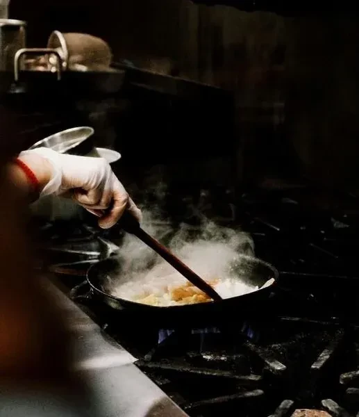 Gloved hand stirs food in a black pan on a gas stove.