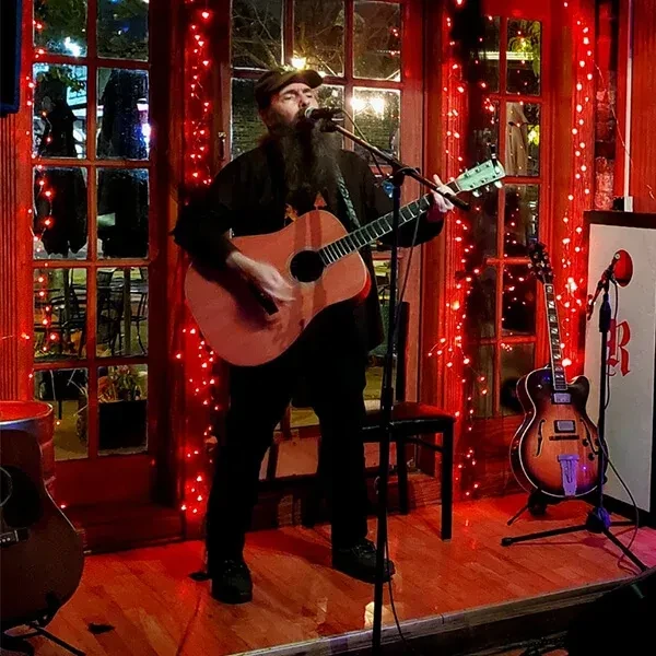 Musician with dark beard playing guitar in a red-lit venue.