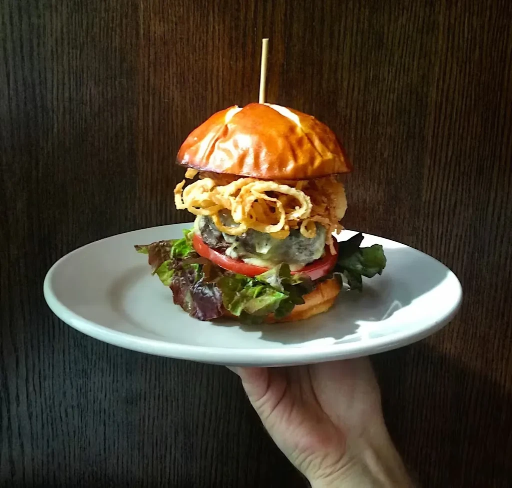 Hand holding a plate with a gourmet burger topped with onion rings.