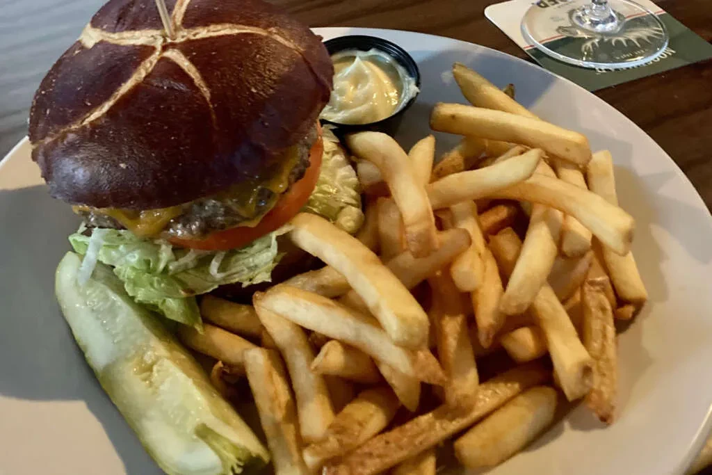 Gourmet burger with pretzel bun, fries, dip, and pickle on white plate.