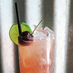 Pink cocktail in a textured glass with lime, cherries, and straw.