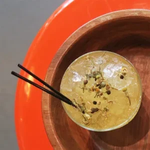 Pale yellow cocktail with botanicals, ice, and straws in a wood bowl.