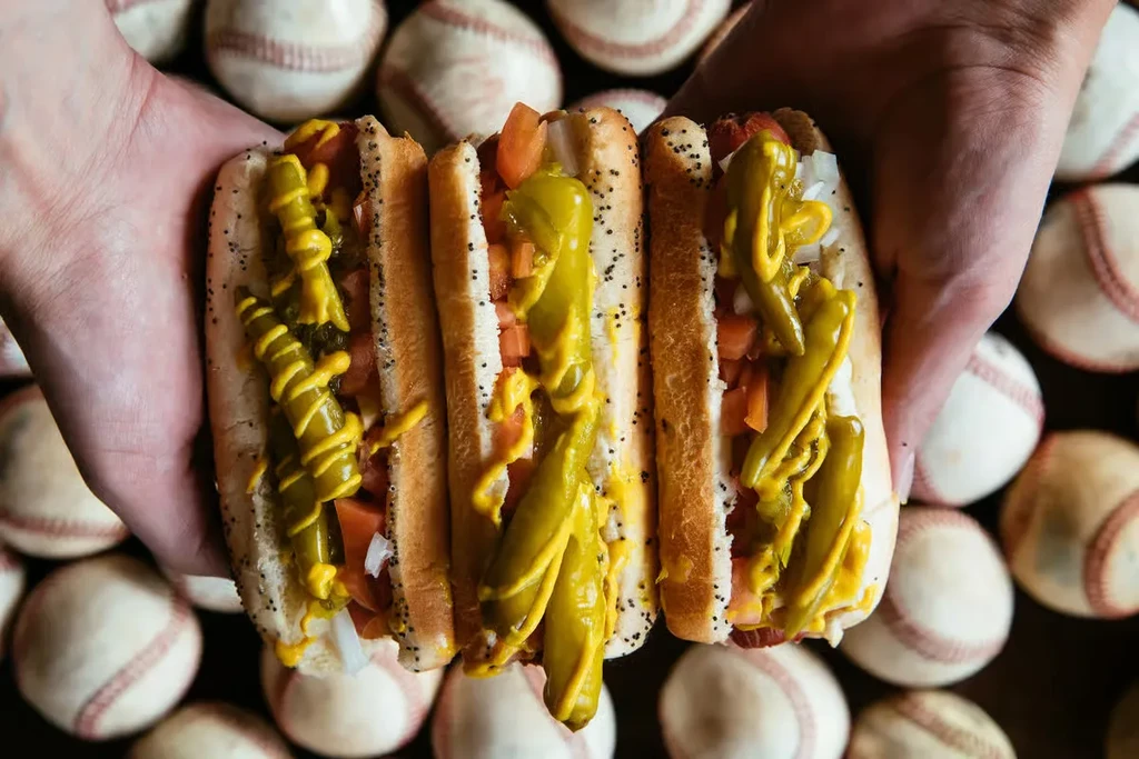 Hands holding three hot dogs with mustard, peppers, tomatoes, onions.