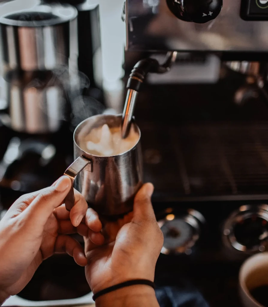 Barista's hands frothing milk in a steel pitcher under a steam wand.
