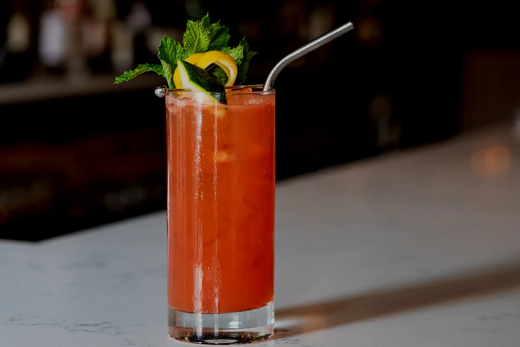 Red-orange cocktail in a tall glass, garnished with mint, lemon, and green fruit.