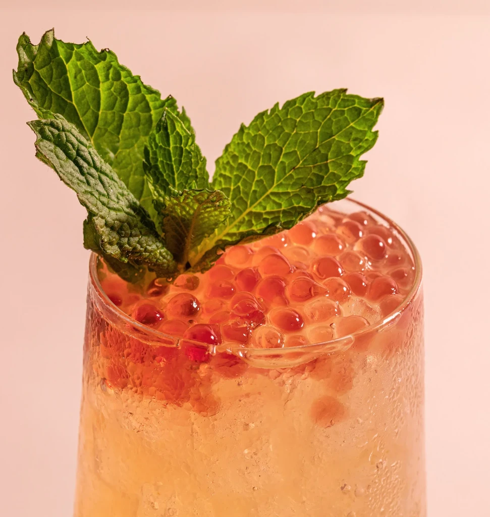 Cocktail with amber liquid, ice, reddish-orange pearls, and mint.