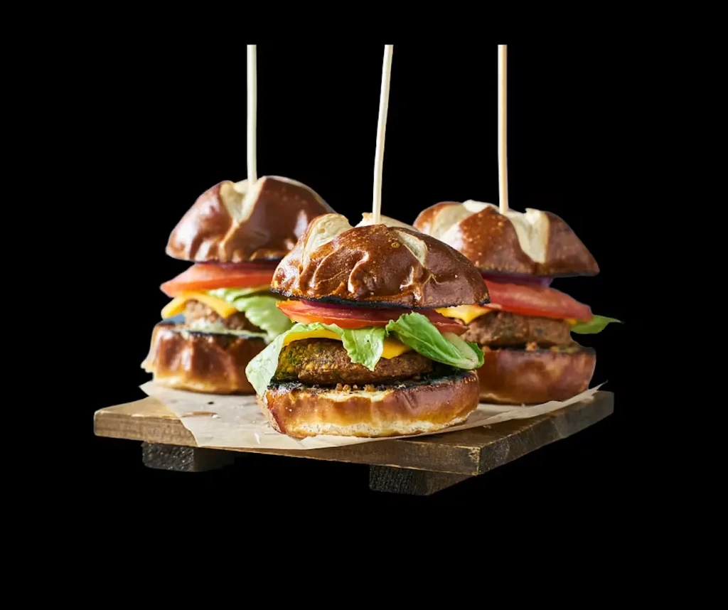 Three mini sliders with pretzel buns, cheese, tomato, and lettuce.