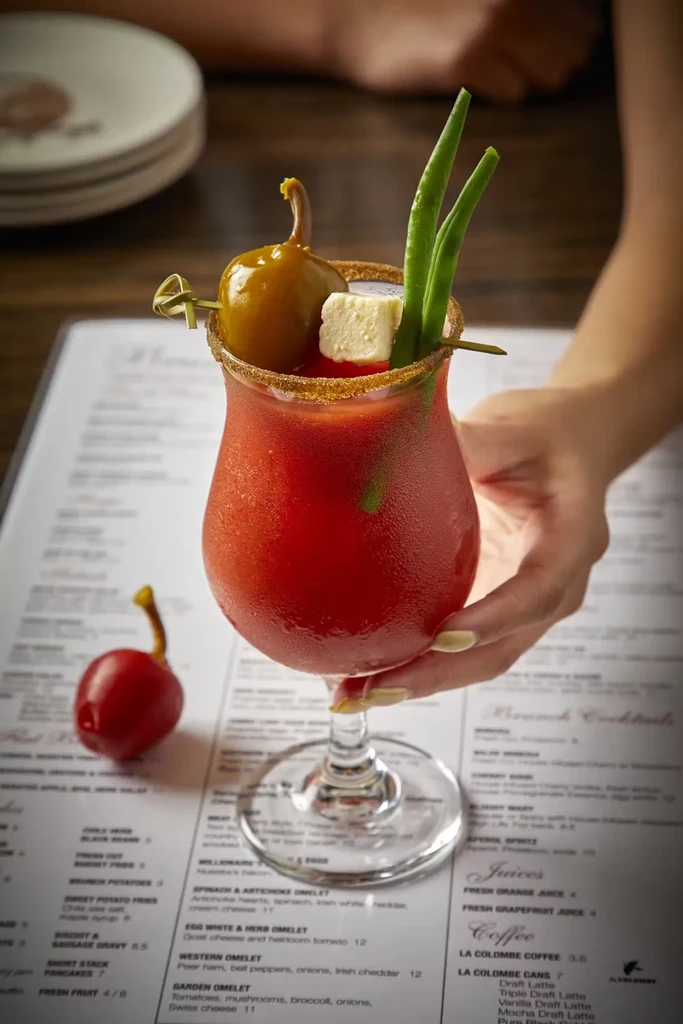 Hand holding Bloody Mary garnished with pepper, cheese, and beans.