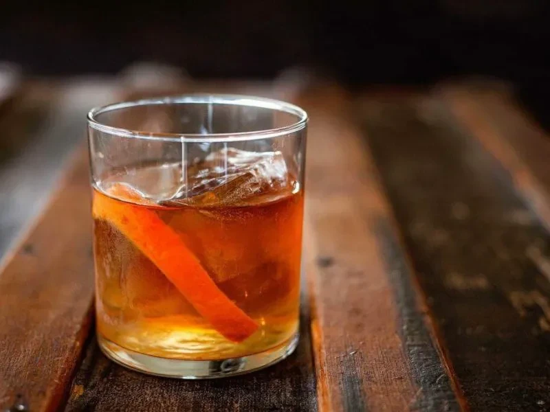Whiskey cocktail with orange peel garnish on a rustic wooden surface.