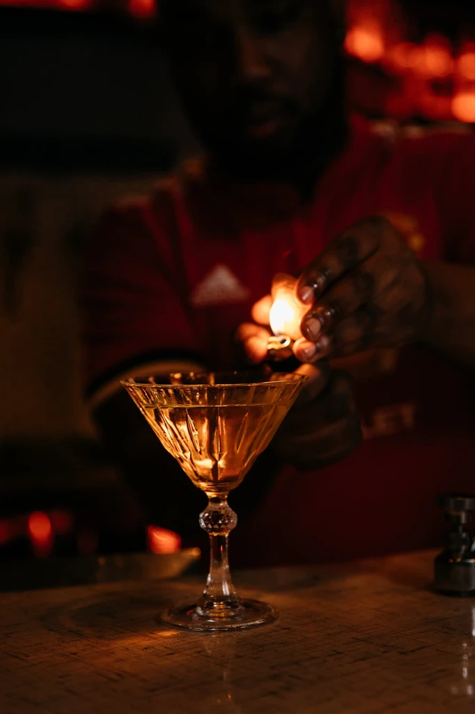 Bartender ignites a garnish over an amber cocktail in a martini glass.