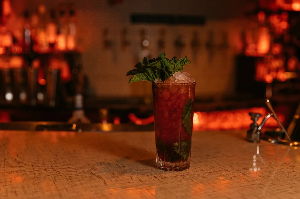 Reddish-brown cocktail with crushed ice and mint sprig on a bar.