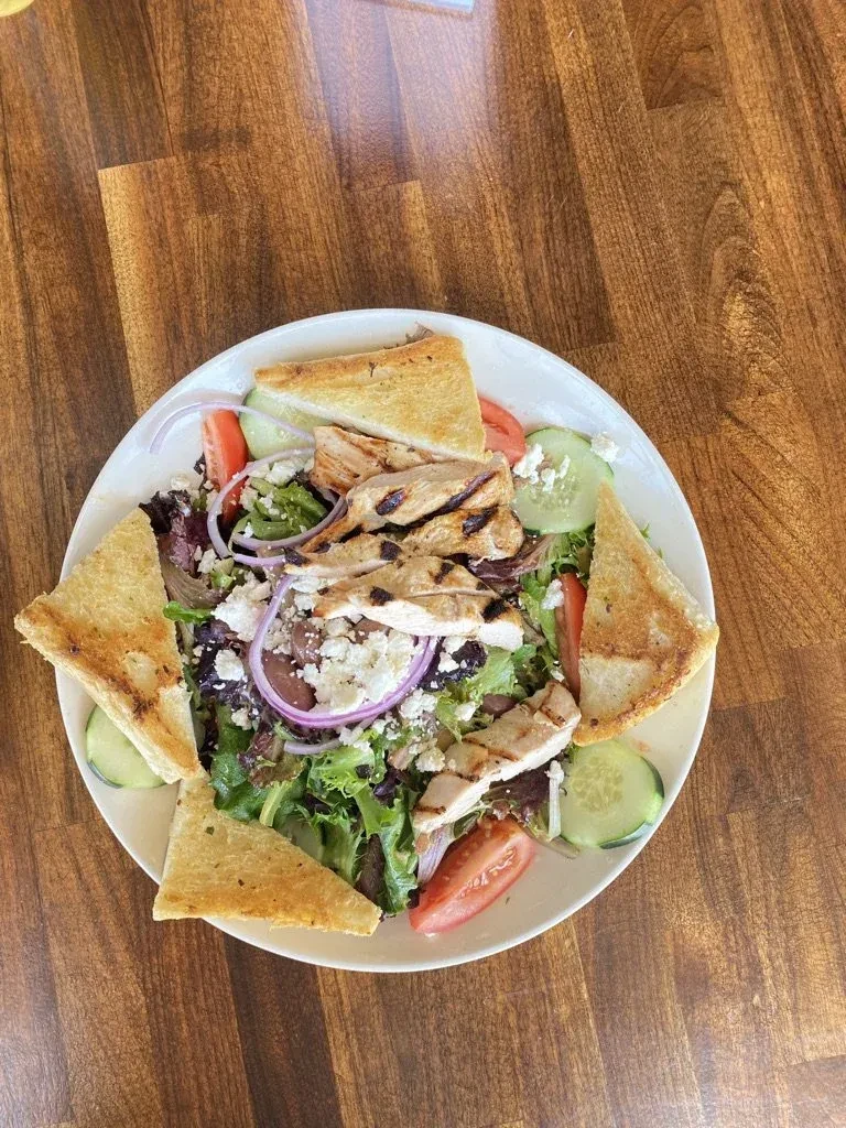 Top-down grilled chicken salad, vegetables, feta, and bread on wood table.