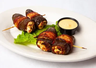 Four skewered bacon-wrapped appetizers with creamy filling, lettuce, and dipping sauce.