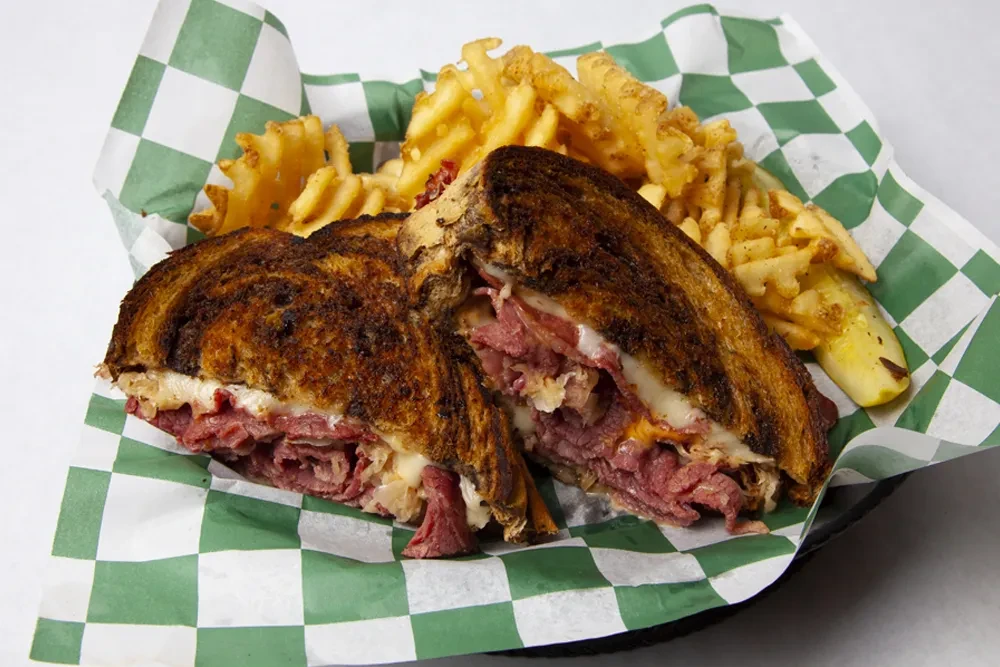 Reuben sandwich halves, waffle fries, and pickle in a black basket.